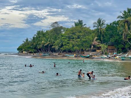 People playing and swimming in the ocean near a coastline lined with palm trees and colorful fishing boats.