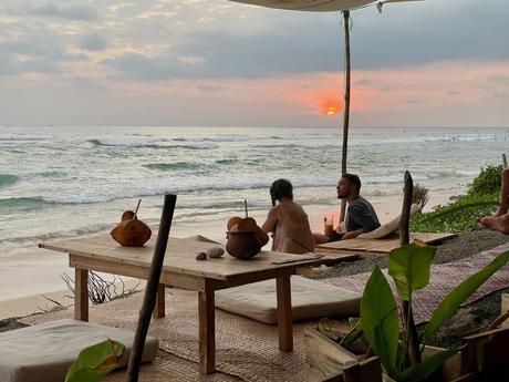 A man and a woman sitting on cushions at a beachside café, sipping drinks from coconuts while watching the sunset over the ocean. The scene is relaxed and serene, with waves crashing softly in the background.