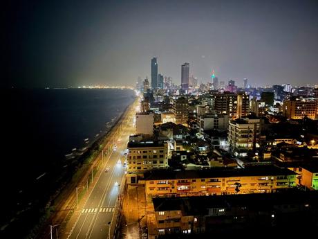 Aerial view of Colombo city at night, with the brightly lit coastline stretching into the distance. Tall skyscrapers are illuminated against the dark sky, and the ocean shimmers beside the city.