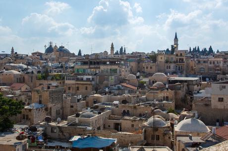 A view of the Old City rooftops in Jerusalem, showing a cluster of ancient buildings with domed roofs, churches, and a tower under a bright, partially cloudy sky.