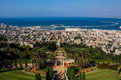 A panoramic view of Haifa, Israel, showcasing the Baha'i Gardens with its iconic golden dome and well-manicured greenery. The city and Mediterranean Sea extend into the background.