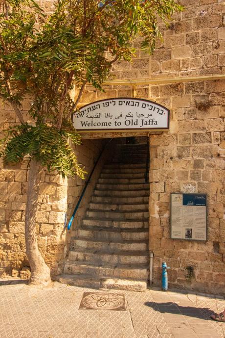 A staircase leading into Old Jaffa, with a welcoming sign in Hebrew, Arabic, and English, set against the weathered stone walls and shaded by a leafy tree.