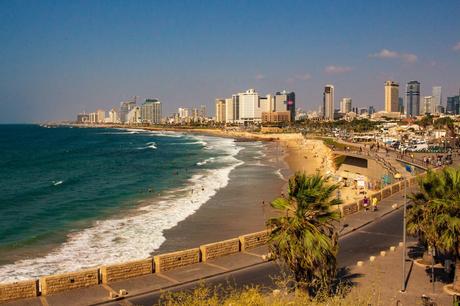 The coastline of Tel Aviv, Israel, stretching into the distance. The beach is dotted with swimmers, and the skyline is filled with tall, modern buildings under a bright blue sky.