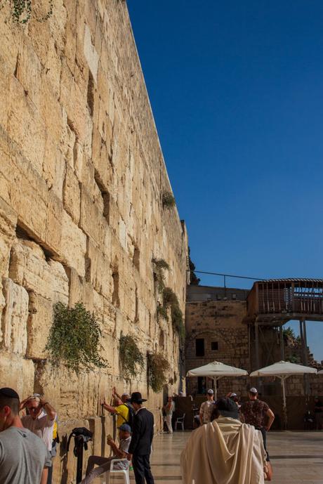 Side angle of the Western Wall in Jerusalem, with several people praying against the wall. The angle captures the towering height of the ancient limestone structure.