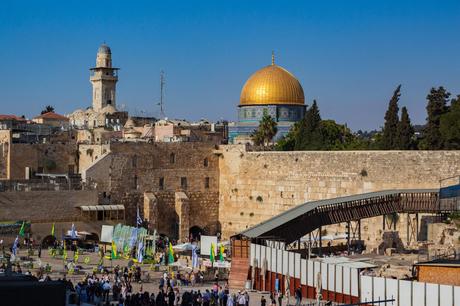 The Western Wall and the Dome of the Rock in Jerusalem, with people gathered in prayer at the base of the wall, and the bright golden dome shining against a clear blue sky.