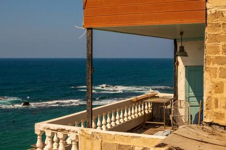 A seaside balcony overlooking the Mediterranean Sea, with turquoise waters and waves crashing against rocks, framed by an outdoor seating area and a simple stone building.