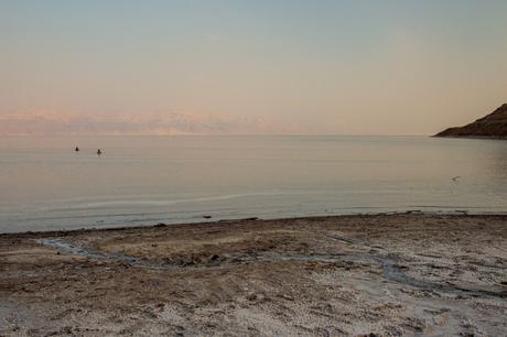 A tranquil view of the Dead Sea at sunset, with a calm shoreline in the foreground and two distant people floating in the water. The horizon shows a faint outline of mountains shrouded in a soft pink glow.