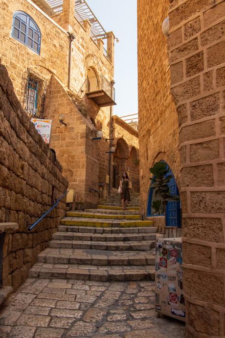 A cobblestone alley in Jaffa, Israel, lined with stone walls and buildings. A woman walks up the narrow staircase, surrounded by arches and balconies with blue window frames.