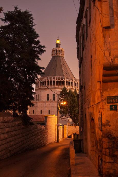 The Church of the Annunciation in Nazareth, Israel, at dusk. The towering dome is softly illuminated against the fading purple sky, with narrow streets and stone buildings leading up to it.