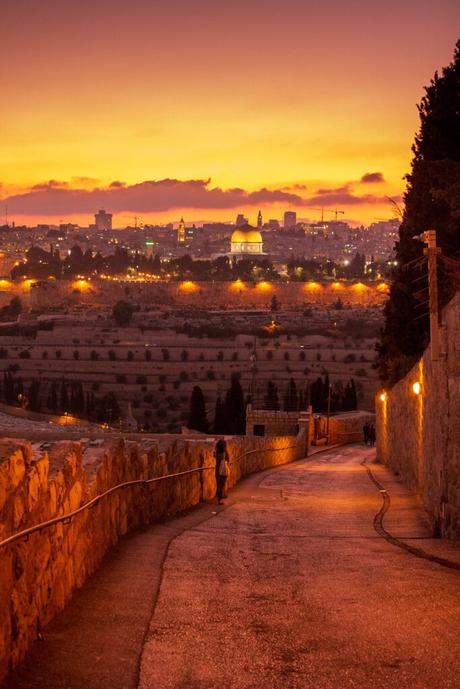 Evening shot of the Mount of Olives pathway, with a view overlooking Jerusalem’s Old City and the Dome of the Rock glowing under a golden sunset. Street lamps line the curved pathway.