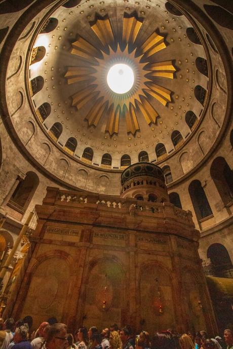 The domed ceiling of the Church of the Holy Sepulchre in Jerusalem, Israel, with golden light streaming down. Below, visitors gather around the shrine beneath the large, ornate ceiling.