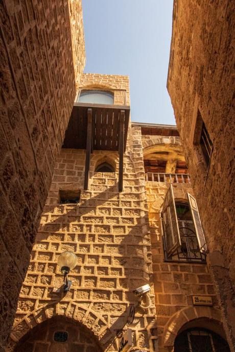 A narrow alley with tall stone buildings in Jaffa, Israel. The view looks upward to an open window and a balcony, with bright sunlight casting shadows on the textured walls.