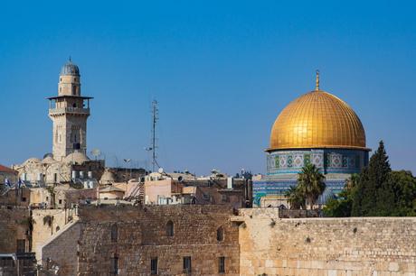 The Dome of the Rock in Jerusalem, Israel, with its gleaming golden dome standing out against a clear blue sky. Surrounding the dome are stone walls and ancient buildings, including a tall minaret.