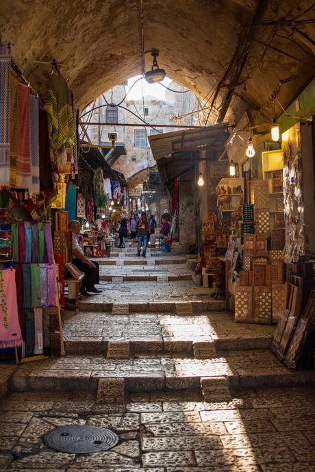 A narrow marketplace in Jerusalem's Old City, with arched stone ceilings, colorful textiles, and stalls selling crafts, with light shining down through the archway.