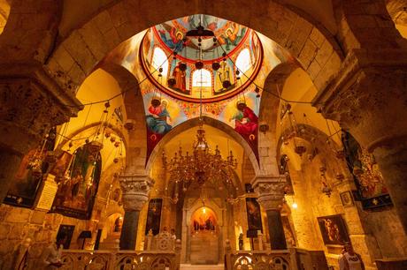 The richly decorated interior of an Orthodox church in Jerusalem, Israel, with vibrant iconography on the dome, gold chandeliers, and arched stone walls creating a warm, glowing atmosphere.