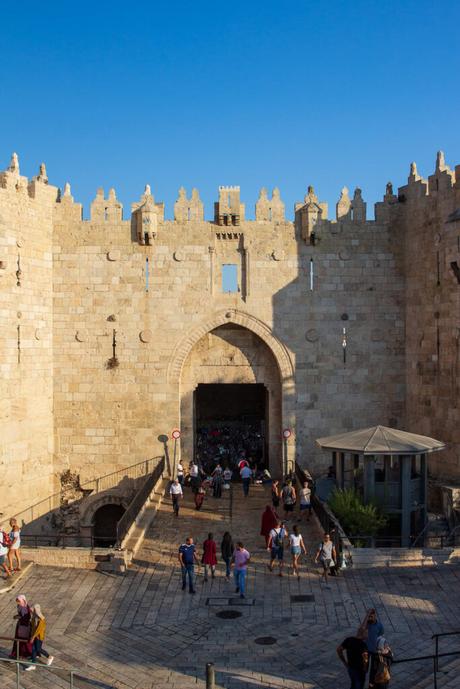 View of the Damascus Gate in the Old City of Jerusalem, with its large stone archway and battlement-style walls. People are walking through the entrance and around the plaza in front.