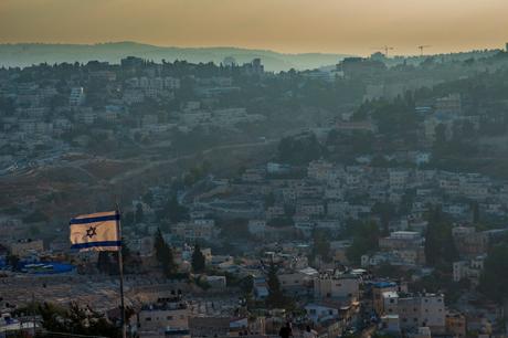 An Israeli flag flying at the top of a hill, overlooking a densely packed hillside of buildings in the early evening. The surrounding area is shrouded in a blue mist as the sun sets.