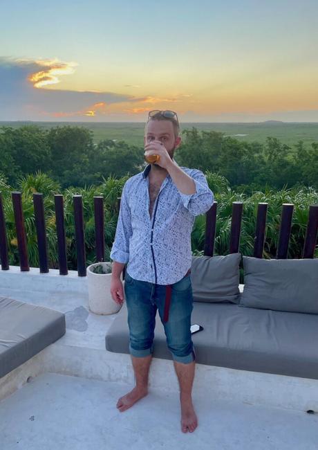 Alex Tiffany standing on a terrace during sunset, holding a drink to his mouth while gazing into the distance. He’s casually dressed with rolled-up jeans, and the background features lush greenery and an expansive view of nature with an orange-hued sky.