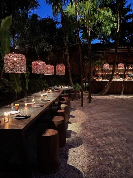 A luxury outdoor dining area in Tulum at night with a long wooden table set with candles, plates, and glasses. Overhead, woven basket-style light fixtures hang, illuminating the space surrounded by palm trees, giving the setting a tropical, intimate atmosphere.