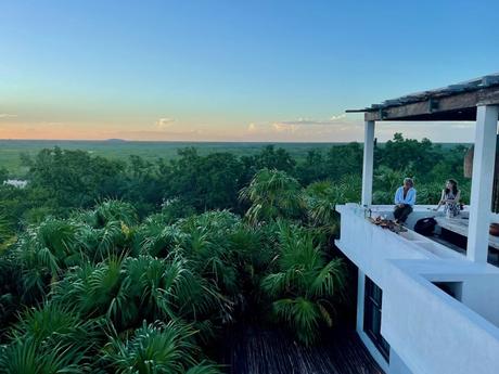 A rooftop terrace with a scenic view of lush green treetops extending towards the horizon, where the sky transitions from light blue to golden as the sun sets. A couple of people are seated on the terrace, talking and enjoying the peaceful surroundings.