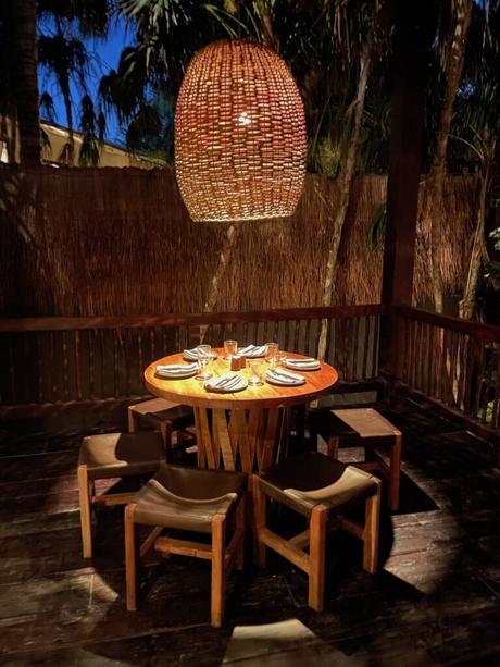 A round wooden table set for dinner with dimly lit lanterns above. The surrounding environment is filled with palm trees and a rustic ambiance.