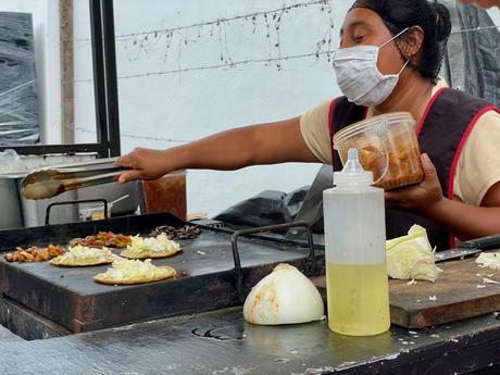 A street vendor in Tulum preparing food at a grill, wearing a white mask and black apron. She is holding a container of what appears to be marinated food and cooking tacos or tortillas with toppings, with a large onion and a bottle of oil on the side.