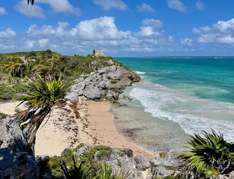 A stunning coastal scene with turquoise ocean waves gently crashing onto a small sandy beach next to the ancient Tulum ruins. The shoreline is bordered by rocky cliffs and palm trees, with a stone ruin visible in the distance under a partly cloudy sky.