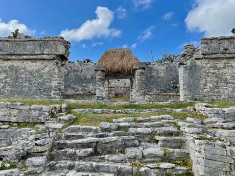 A stone structure from the Tulum ruins, featuring a small thatched roof at the entrance and weathered stone pillars. The ancient building is surrounded by stone steps leading up to the entrance, set against a backdrop of a bright blue sky with fluffy clouds.