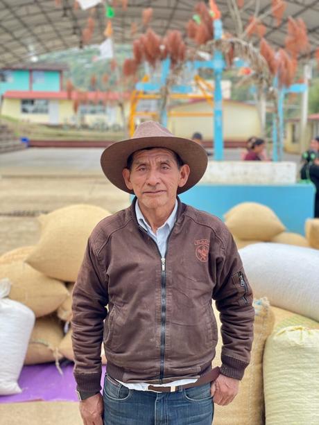 A man wearing a brown jacket and wide-brimmed hat standing in front of stacked burlap sacks in an outdoor area with a large, colorful structure in the background.