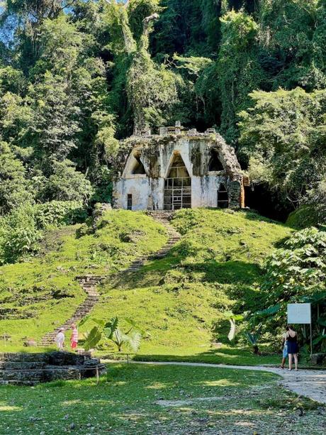 A white, vine-covered, ancient temple structure on a grassy hill with a stone stairway, nestled in the jungle.