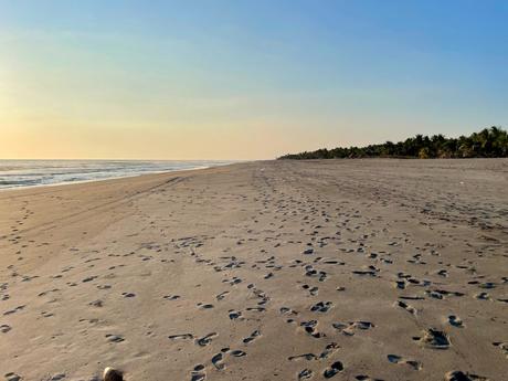 A long stretch of sandy beach dotted with footprints, extending towards the horizon under a soft, pale blue sky.