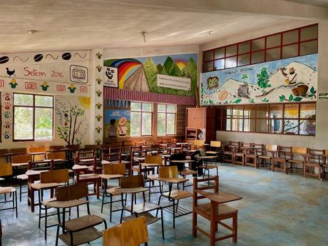 An empty classroom with wooden desks and chairs arranged in rows. The walls are adorned with colorful murals depicting local culture, agriculture, and messages about organic coffee and respect for nature.