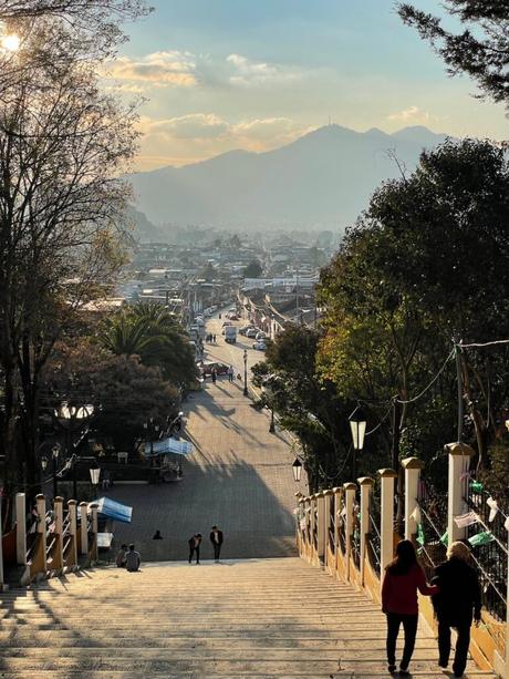 A long, wide staircase descending into the streets of San Cristobal, with mountains in the background and people walking down the steps under the late afternoon sun.