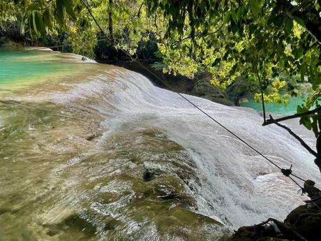 A gentle waterfall flows over smooth rock formations, with clear, turquoise water in the background, partially shaded by lush green leaves.