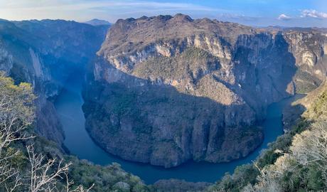 A panoramic view of a deep canyon called Sumidero Canyon in Chiapas with steep cliffs covered in vegetation. A calm river snakes through the bottom of the canyon, casting large shadows on the surrounding rock formations.