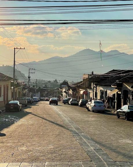 A broad street lined with parked cars and traditional houses, with power lines crossing overhead, and mountains looming in the background during sunset.