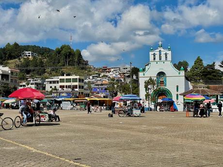 A vibrant market square in front of the white and green church of San Juan Chamula with vendors under colorful umbrellas and people walking around, with green hills in the background.