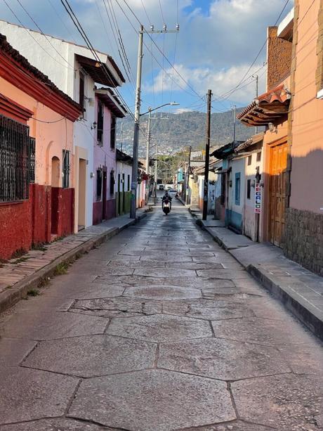 A narrow alley lined with colorful houses, with a motorbike rider approaching in the distance and mountains visible at the far end.