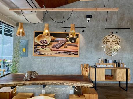 A cozy chocolate shop with wooden tables and chairs, featuring hanging lamps and a wall display of chocolate bars and cacao beans.