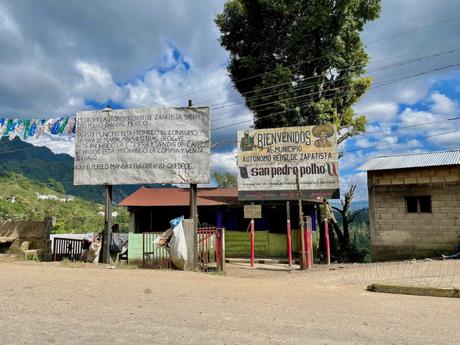 A welcome sign at the entrance of a Zapatista rebel municipality in San Pedro Polho, Chiapas. The sign contains messages about prohibitions in the community, and behind it, small buildings and mountainous terrain can be seen.