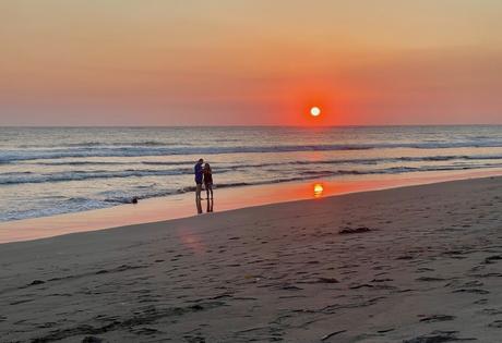 A couple standing on the shore of a beach in Chiapas at sunset, with the glowing orange sun reflecting off the water and soft waves rolling onto the sand.