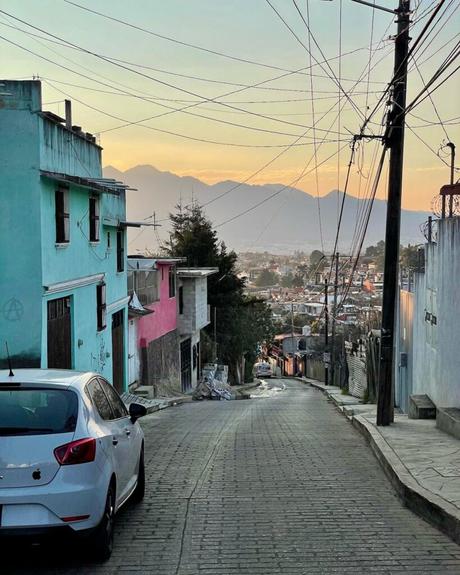 A quiet downhill street lined with colorful buildings, power lines overhead, and mountains visible in the background under a fading sunset.