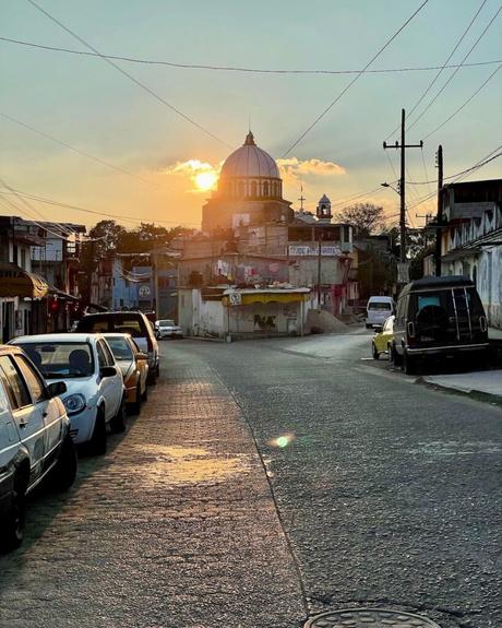 A street in the historical centre of San Cristobal de las Casas at sunset, with a church dome in the distance and parked cars lining the cobblestone road.