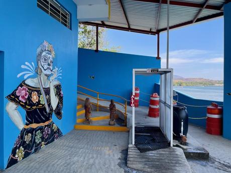 A colorful mural painted on a blue wall featuring a woman in traditional Mexican dress with her face painted as a skull, in the style of Día de los Muertos. The area includes some statues, red containers, and a view of water in the background.