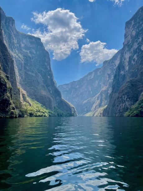 A stunning view of the deep river gorge inside Sumidero Canyon with towering cliffs on both sides. The water below reflects the blue sky and clouds, creating a serene and majestic scene.