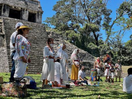 A group of people in traditional white Mexican clothing participate in a ceremony near ancient ruins, with a pyramid in the background.