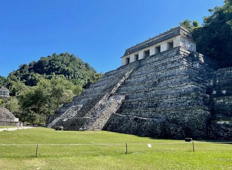 A large stone pyramid with steep steps at Palenque ruins, surrounded by green grass and trees under a clear blue sky.