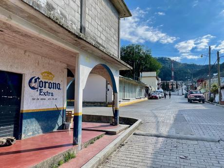 A quiet street scene with a Corona Extra advertisement on the side of a building, and a couple of cars parked further down the road under a bright blue sky.