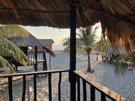 View from the thatched porch of a simple beach hut in Chiapas with palm trees and other huts on the sand and the ocean beyond all bathed in the soft light of sunset.