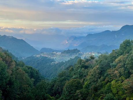 A lush, forested valley surrounded by rolling mountains under a partially cloudy sky, with a small village visible in the distance.